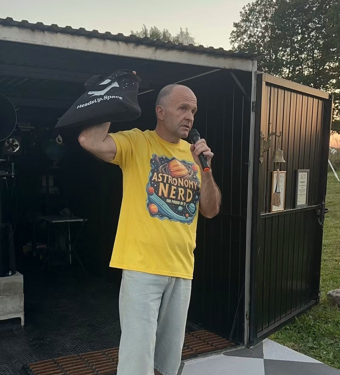 A man wearing a bright yellow graphic t-shirt and light gray shorts stands in front of a dark metal shed, holding a black bag over his shoulder and a handheld microphone. The background features a checkered gray and white tiled floor, grass, and trees under a soft sky.