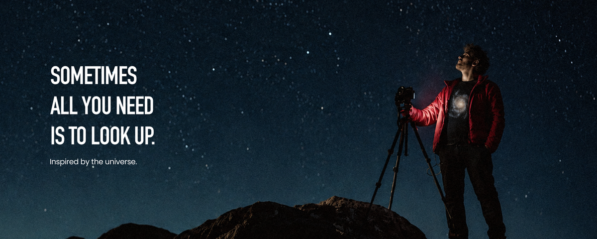 A man in a red jacket gazes up at a star-filled night sky beside his camera and tripod on a rocky hill. He wears a black galaxy T-shirt. White text reads: “Sometimes all you need is to look up. Inspired by the universe.”